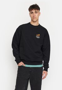 Zwarte sweatshirt met lange mouwen, ribgebreide boorden en onderrand. Heeft een klein oranje en geel logo op de borst. Draagt over een lichtblauw shirt.