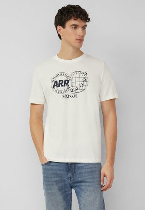 Młody mężczyzna w białej koszulce z grafiką globu i gwiazd, w niebieskich jeansach, stoi na tle jednolitego szarego tła.