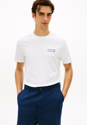 Junger Mann trägt ein weißes Tommy Hilfiger T-Shirt und marineblaue Hose, steht mit den Händen in den Taschen vor einem einfarbigen Hintergrund.