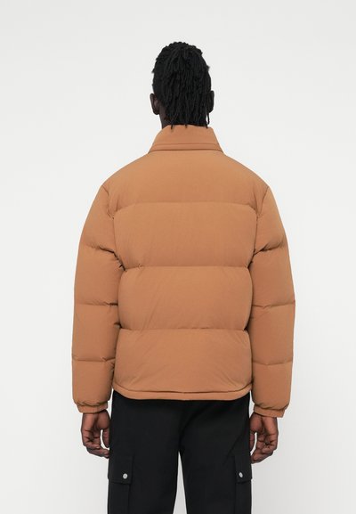 Veste matelassée marron avec sections quiltées horizontales, col haut et poignets élastiques, associée à un pantalon cargo noir. Tissu texturé.