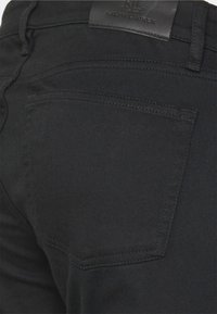 Schwarze Denim-Jeans mit einem standardmäßigen Design, einer Gesäßtasche und einem Lederetikett am Bund. Glatte Textur, klassischer Schnitt.