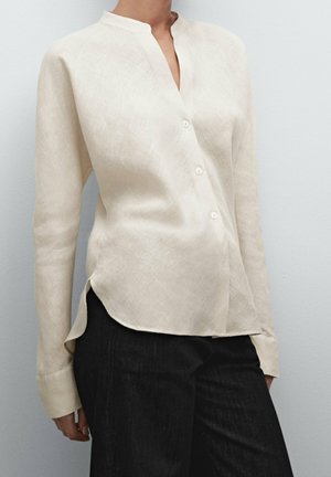 Woman wearing a cream linen long-sleeve button-up shirt with a mandarin collar and black pants against a plain gray background.