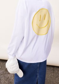 Persona che indossa una camicia bianca a maniche lunghe con un grande volto sorridente giallo sulla schiena, tiene un cappellino bianco e una gonna di jeans blu all'aperto.
