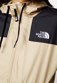 Veste North Face beige et noire avec fermeture éclair à l'avant, capuche et cordons noirs, arborant le logo de la marque sur la poitrine.