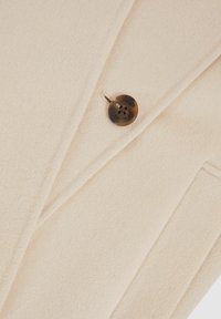 Béžový vlněný kabát s jemnou, texturovanou tkaninou. Má jeden hnědý knoflík a elegantní design klopu s boční kapsou.