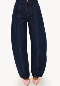 Vaqueros de mezclilla azul oscuro de talle alto con piernas anchas, que presentan costuras prominentes y un cierre de botones, combinados con tacones puntiagudos rosa claro.