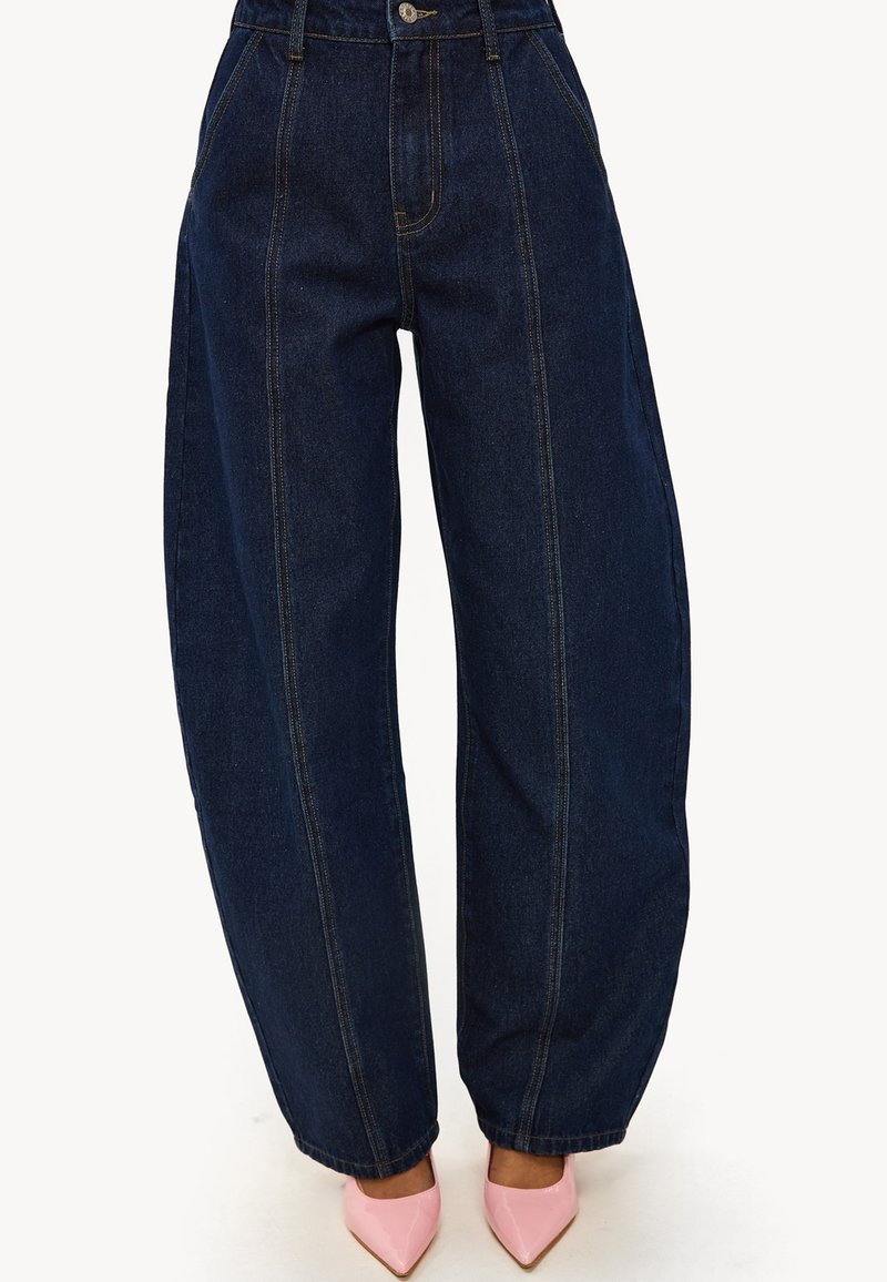 Vaqueros de mezclilla azul oscuro de talle alto con piernas anchas, que presentan costuras prominentes y un cierre de botones, combinados con tacones puntiagudos rosa claro.