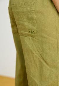 Olive green pants with a textured fabric. Features a patch pocket with a small, embroidered heart logo in matching color.