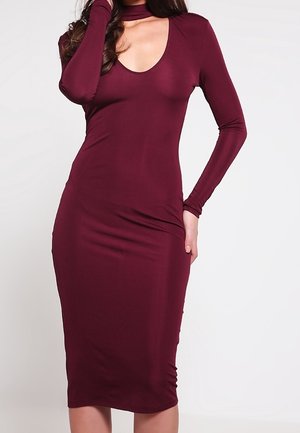 Mujer vistiendo un vestido midi burdeos ajustado de manga larga con escote pronunciado y un pequeño corte cerca de la clavícula.