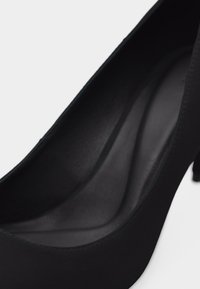 Μαύρο παπούτσι με ψηλό τακούνι, ματ απαλής υφής, στρογγυλεμένη μύτη και καμπυλωτή εσωτερική σόλα, πάνω σε απλό λευκό φόντο.