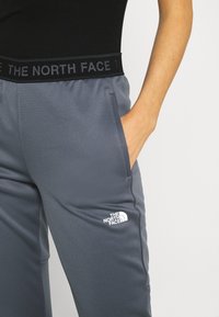 Osoba v šedých kalhotách The North Face se bočním kapsou, ruka v kapse a černém topu s logem The North Face na pásu.
