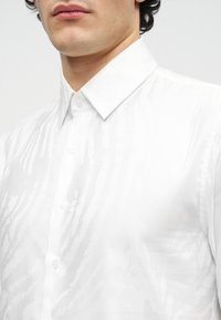 Muž nosiaci priliehavú bielu košeľu s jemným vzorom pripomínajúcim zebru a špicatým golierom, viditeľné pery a brada.