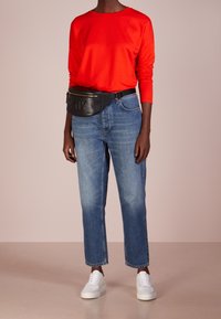 Czerwona koszula z długim rękawem, niebieskie jeansy o prostych nogawkach, białe trampki oraz czarna nerka z złotym suwakiem i tłoczonym logo.