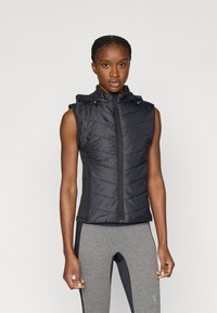 Veste sans manches noire matelassée avec une fermeture éclair à l'avant, une capuche et un motif matelassé en diagonale ; associée à des leggings ajustés gris.
