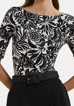 Femme portant un haut à motif floral noir et blanc avec des manches aux coudes, rentré dans un pantalon noir taille haute avec une ceinture tressée.