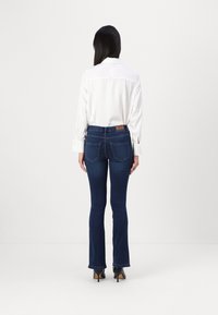Jeans foncés évasés avec une taille haute, associés à une chemise blanche à manches longues avec des détails aux poignets. La vue arrière met en avant le design des poches.