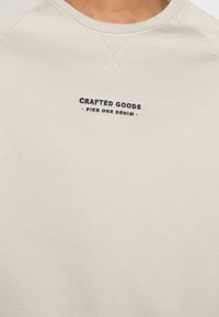 Felpa beige realizzata in morbido tessuto, con collo a giro e un piccolo logo ricamato nero che recita "CRAFTED GOODS - PIER ONE DENIM."