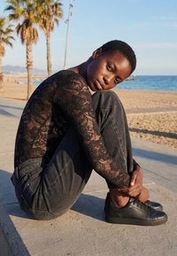 Jeune personne aux cheveux courts, portant un haut en dentelle noire et un jean foncé, assise en serrant ses genoux sur un rebord en béton au bord d’une plage ensoleillée avec des palmiers.