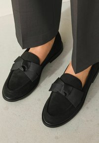 Schwarze Slipper aus weichem Wildleder, mit einer großen Schleifenapplikation und einer flachen Gummisohle. Kombiniert mit dunklen karierten Hosen.