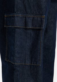 Tmavé riflové cargo nohavice s veľkou bočnou kapsou, oranžovými švami a textúrovanou látkou. Dizajn má rovný strih.