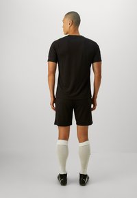 Camiseta deportiva negra y pantalones cortos, material sintético, corte holgado. Calcetines blancos hasta la rodilla, botas negras, vista desde la parte de atrás.