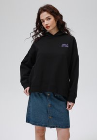 Sudadera negra con capucha con el texto púrpura "¡MEJOR DÍA EVER!", combinada con una falda de mezclilla azul con cierre de botones en la parte delantera y un dobladillo deshilachado.