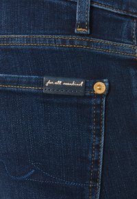 Mörkblå denimjeans med en bakficka som har en sydd märkesetikett och en guldig knappaccent. Synlig gyllene sömdetalj.