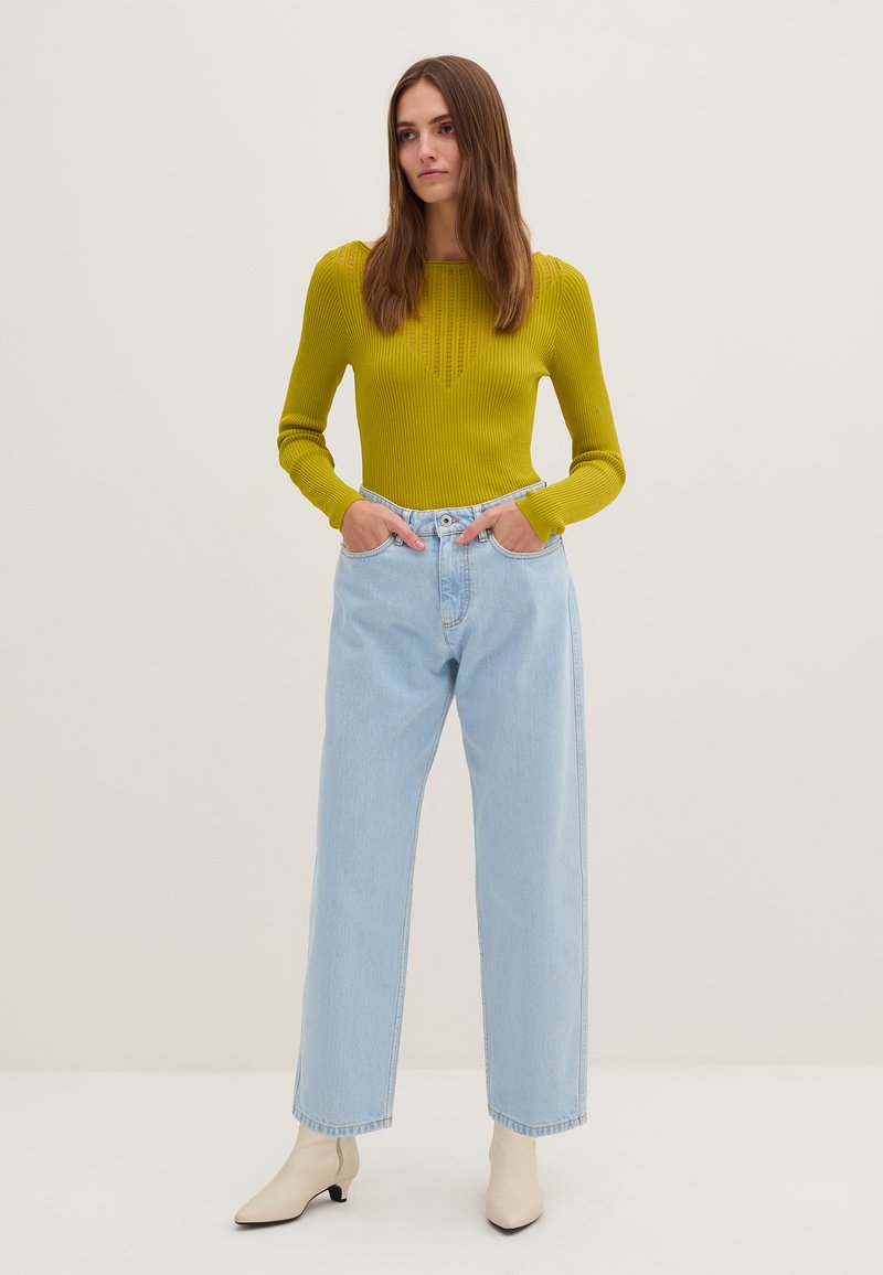 Maglione a coste verde con maniche lunghe, abbinato a jeans a gamba larga azzurri e stivaletti beige, su uno sfondo neutro.