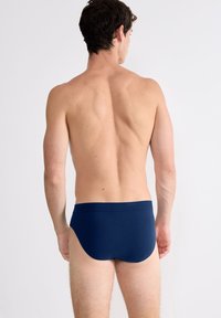 Mann mit kurzem dunklen Haar steht mit dem Rücken zur Kamera, getragen eine marineblaue Unterhose vor einem schlichten hellen Hintergrund.