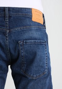 Mörkblå denimjeans med en tanfärgad läderlapp märkt "JACK & JONES" på midjan och en enda bakficka.