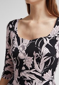 En figurnära klänning med svart tyg prytt med rosa blommönster, en fyrkantig rundringning och trekvartsärmar.