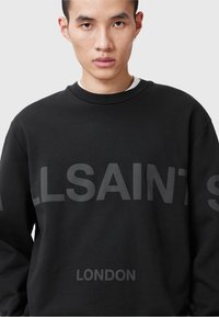 Un jeune homme portant un sweat à capuche noir avec "ALLSAINTS" sur la poitrine et "LONDRES" imprimé en dessous, face à l'objectif sur un fond gris.