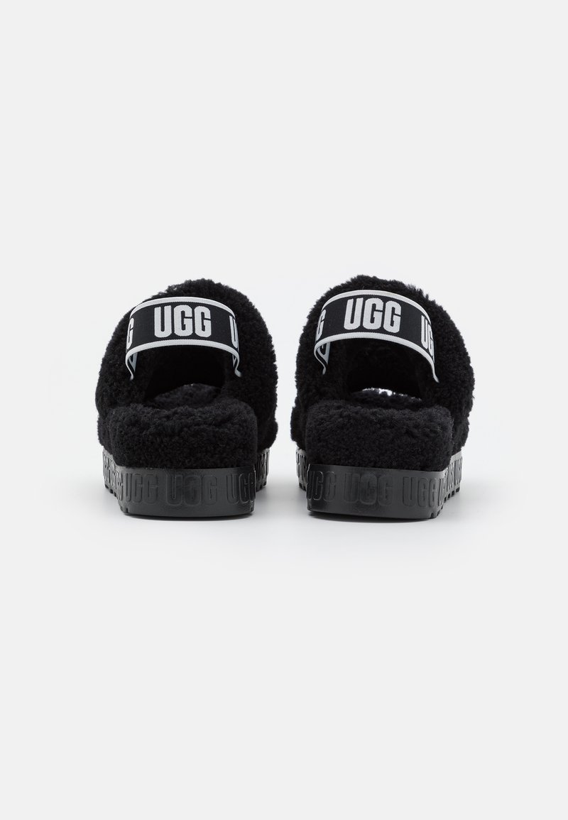 Chanclas de felpa negra con la parte trasera abierta, que cuentan con una suela plana negra grabada con "UGG" y una correa en el talón con la marca.