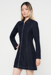 Denimkjole med glidelås foran, lange ermer og krage. Mørkeblått stoff med kontrasterende lyse stingdetaljer.