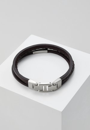 Bracelet en cuir marron foncé avec des lanières tressées et plates et une boucle métallique argentée reposant sur une surface angulaire blanche.
