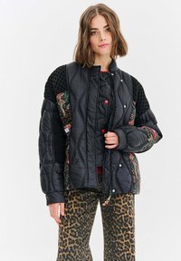 Padded jacket z czarną pikowaną powierzchnią, akcentami w kwiecisty wzór, czerwonymi zatrzaskami i teksturowanymi czarnymi ramionami. W zestawie ze spodniami w lamparta.