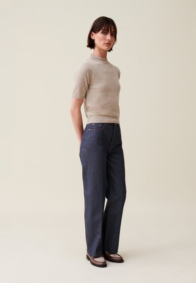 Top in maglia beige a maniche corte abbinato a jeans larghi blu scuro. I jeans presentano tasche classiche e cuciture in contrasto. Scarpe marroni completano l'outfit.