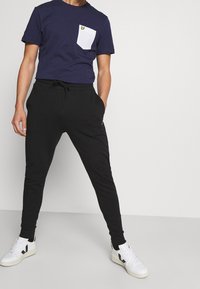 Marineblaues T-Shirt mit weißer Brusttasche und gelbem Logo, kombiniert mit schwarzen, schmal geschnittenen Jogginghosen und weißen Sneakers mit schwarzen Akzenten.