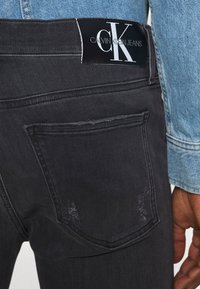 Jeans de denim negro con un ligero desgaste, que presentan una etiqueta de Calvin Klein Jeans en la cintura y bolsillos traseros tradicionales.