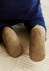 Chaussures à semelle souple marron avec un motif arc-en-ciel texturé ; bordées de matériau côtelé ; contrastant avec un tissu bleu. Placées sur une surface matelassée.