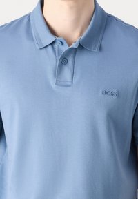 Polo bleu clair avec deux boutons et petit logo "BOSS" brodé sur la poitrine gauche, porté par une personne à la peau claire.