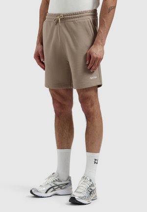 Man wearing beige drawstring shorts, white socks with text, and silver running shoes, standing against a plain background.