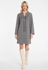 Grijs geruit jurk met lange mouwen, met een strikdetail bij de halslijn. Gecombineerd met beige laarzen tot de knie. Glad textuur.