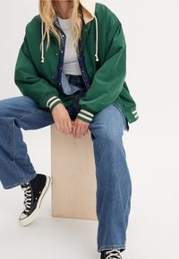 Osoba sedící na dřevěné bedně, oblečená v zelené bundě s bílými pruhy, modrých džínách, černých teniskách a s vrstvami džínové a bílé halenky.