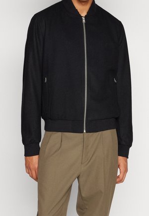 Homme portant une veste noire zippée avec poignets et ourlet côtelés, associée à un pantalon marron ample, debout devant un fond uni.