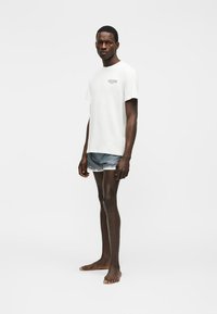 Ung mann som står barbent iført en hvit t-skjorte med trykk og korte denimshorts mot en ensfarget hvit bakgrunn.