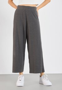 Pantalon gris à jambes larges avec fines rayures verticales, taille élastique, poches latérales, et associé à des baskets blanches sur un fond neutre.