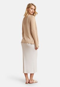 Beige gebreide trui met een opengewerkt ontwerp en een golvende zoom, gecombineerd met een crèmekleurige geribbelde maxirok. Sandalen maken de look compleet.