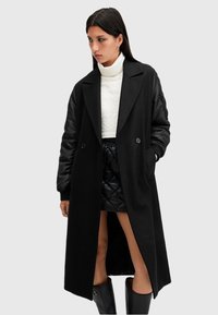 Cappotto nero con un design lungo e dritto, caratterizzato da maniche in tessuto contrastante e due bottoni sul fronte. Abbinare con un top bianco e una gonna lucida.