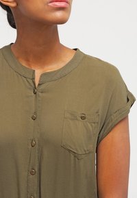 Chemise olive à manches courtes avec un devant à boutons, un col rond et une poche poitrine avec un accent de bouton. Texture de tissu douce.